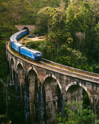 The blue Ceylon train crossing the Nine Arch Bridge in Ella, surrounded by lush green hillside.