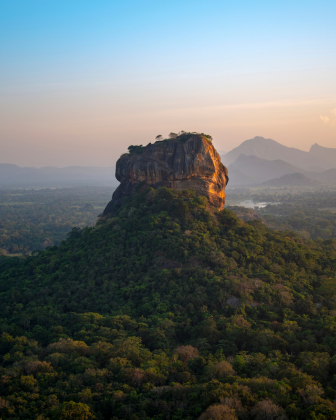 Aerial view of Sigiriya rock fortress rising above the jungle canopy at dawn, distant mountains beyond.