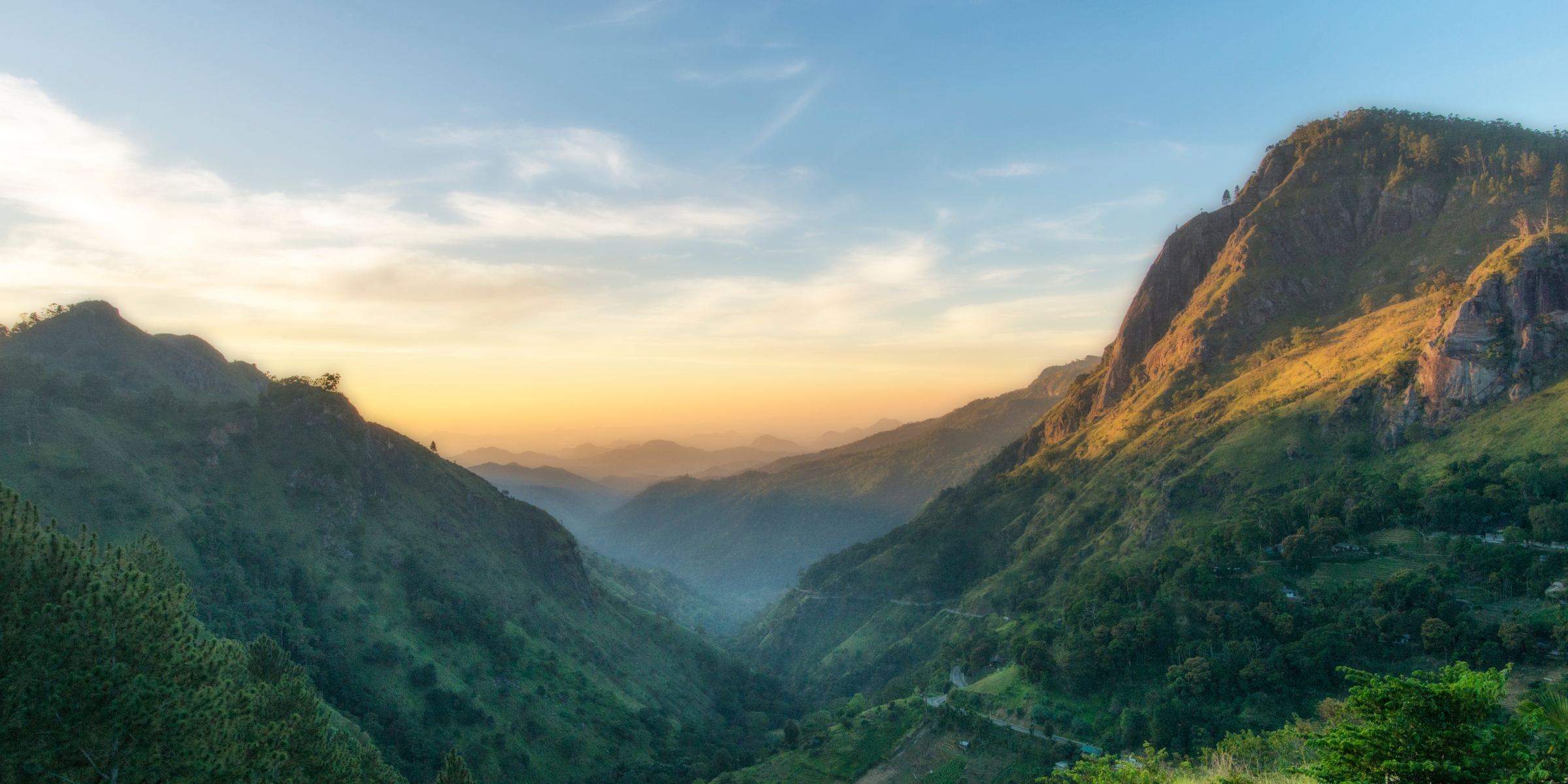 A golden-hour view over a misty Sri Lankan hill country valley, sunlit ridges framing a winding mountain road below.